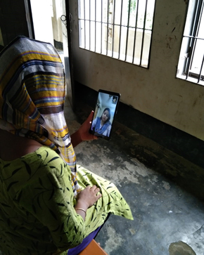 A person wearing a green dress and a scarf is sitting on a chair, looking at a tablet while video calling. The background shows a room with large windows and a concrete floor.