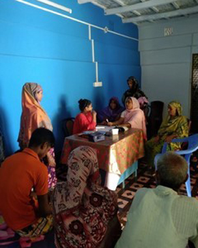 A group of women and men seated in a room, engaged in a discussion or meeting, with some individuals wearing traditional attire. The setting has blue walls, and a table is set up at the center where materials are visible, suggesting a community or support gathering.