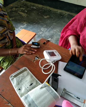 Two individuals are using health monitoring devices on a wooden table, with a pulse oximeter, a blood pressure monitor, and a smartphone placed among notes and papers.