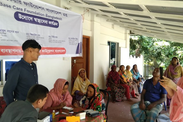 A group of people participating in a health care camp organized by Base Care Foundation Bangladesh, with interactions at a table and several attendees seated in the background.