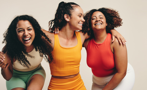 Three women laughing together while wearing colorful athletic outfits, promoting a positive and active lifestyle.