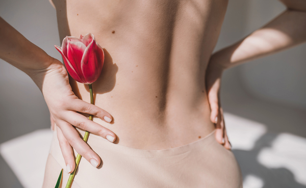 A close-up view of a woman's back, showcasing her smooth skin and a pink tulip held gently against her waist. The soft light highlights the delicate features of the scene, embodying beauty and elegance.