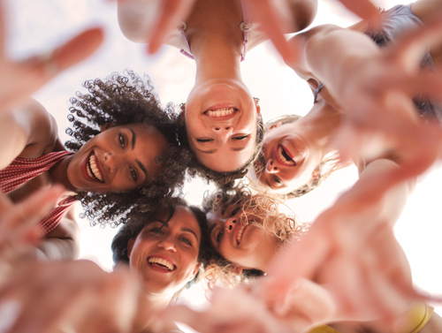 A group of five smiling women in a circle, with their hands reaching towards the camera, expressing joy and friendship.