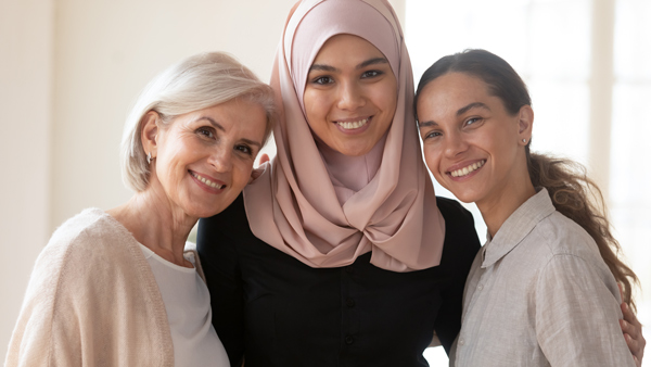 Three smiling women posing together in a well-lit room, showcasing a sense of friendship and diversity, with one wearing a hijab.