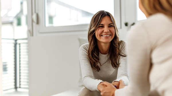 A smiling woman with long, wavy hair is sitting on a sofa, engaged in a conversation with another person, in a bright and airy room with large windows.