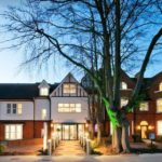 A charming building exterior illuminated at dusk, featuring a mix of traditional and modern architecture, with a large tree in the foreground.